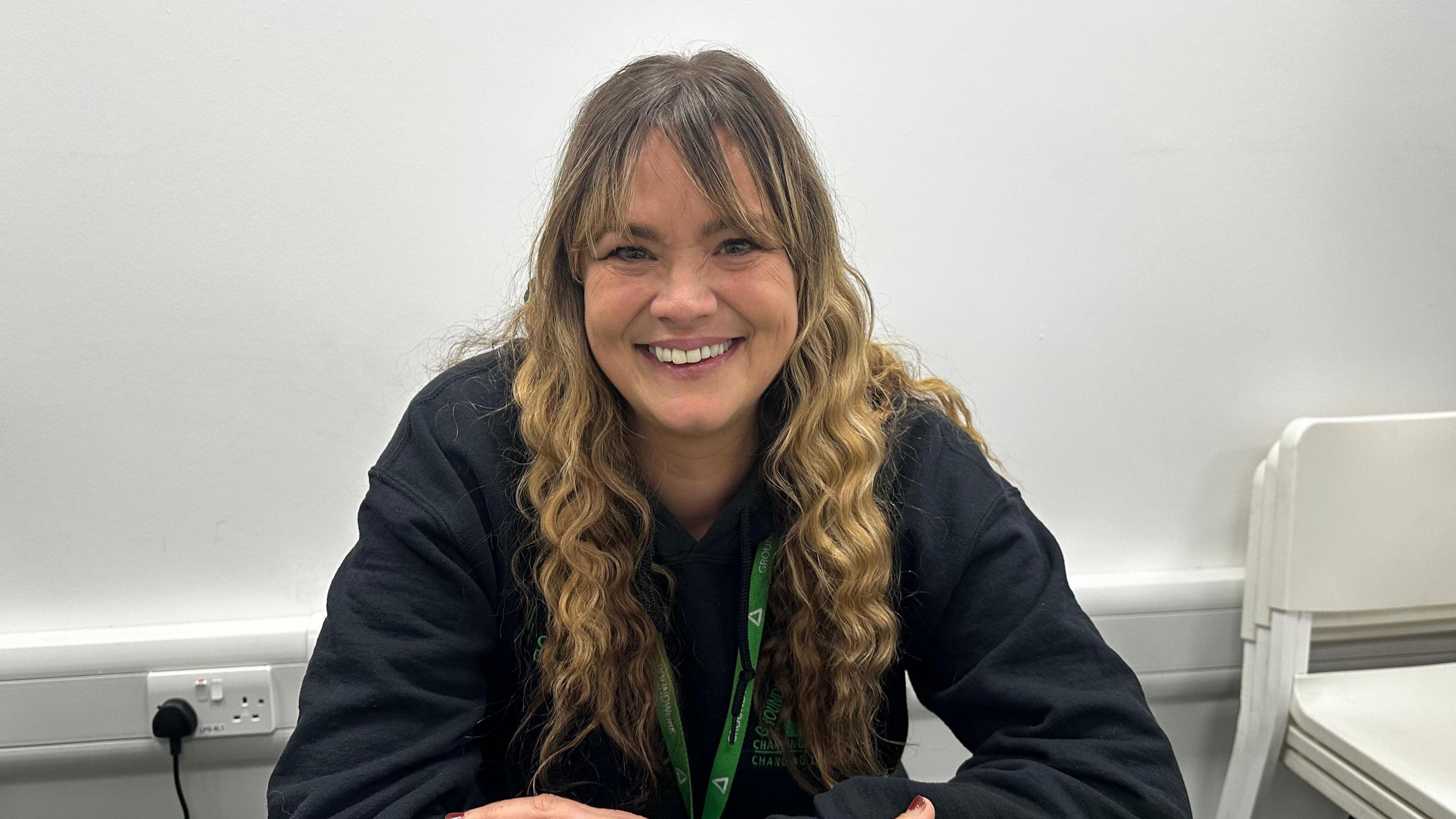 Rachel Denby with long wavy blonde hair wearing a black jacket and green lanyard smiles at the camera in front of a white wall