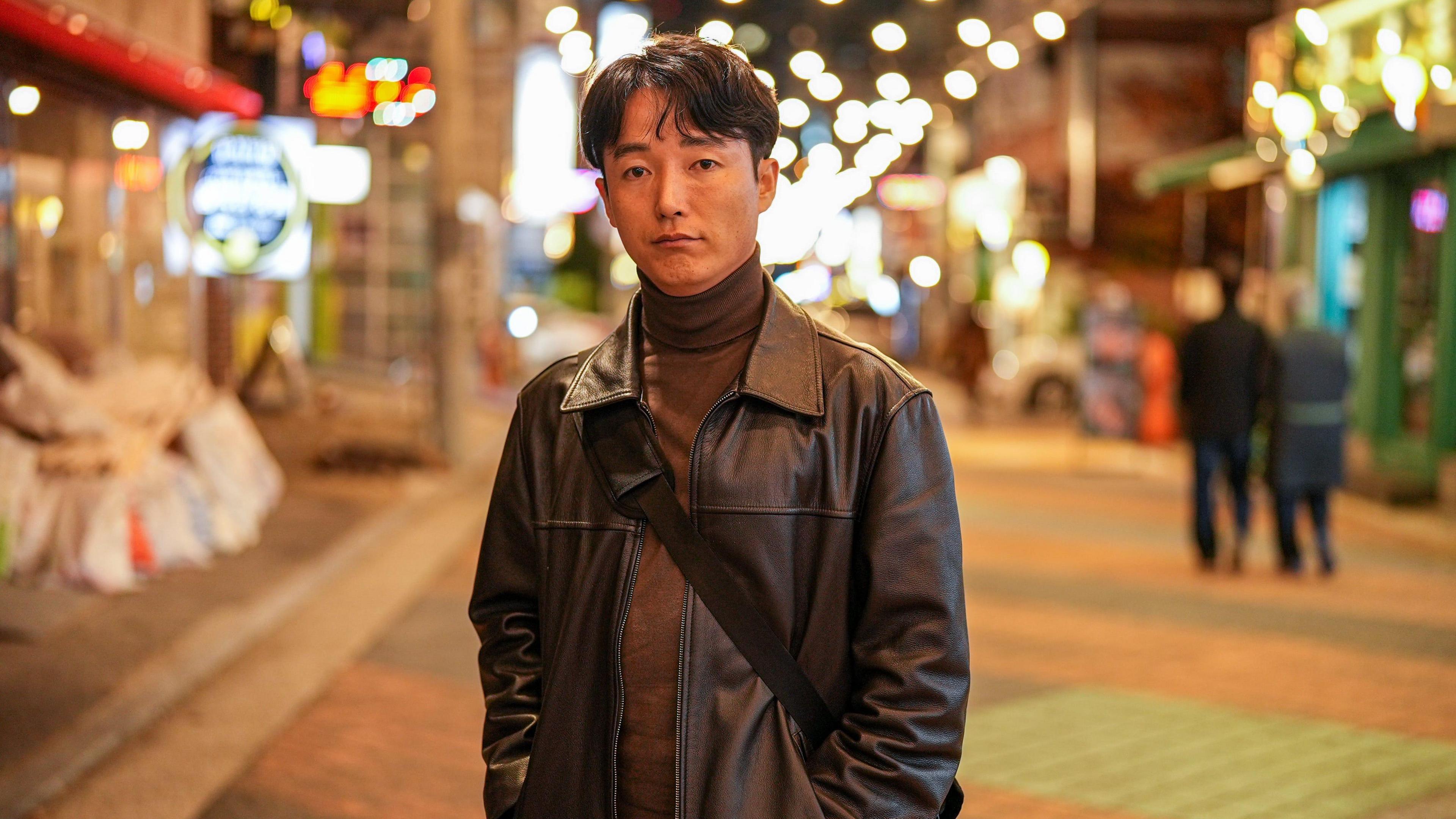 Ryu Seonghyun standing on a street in a brown leather jacket, brown shirt and pants.