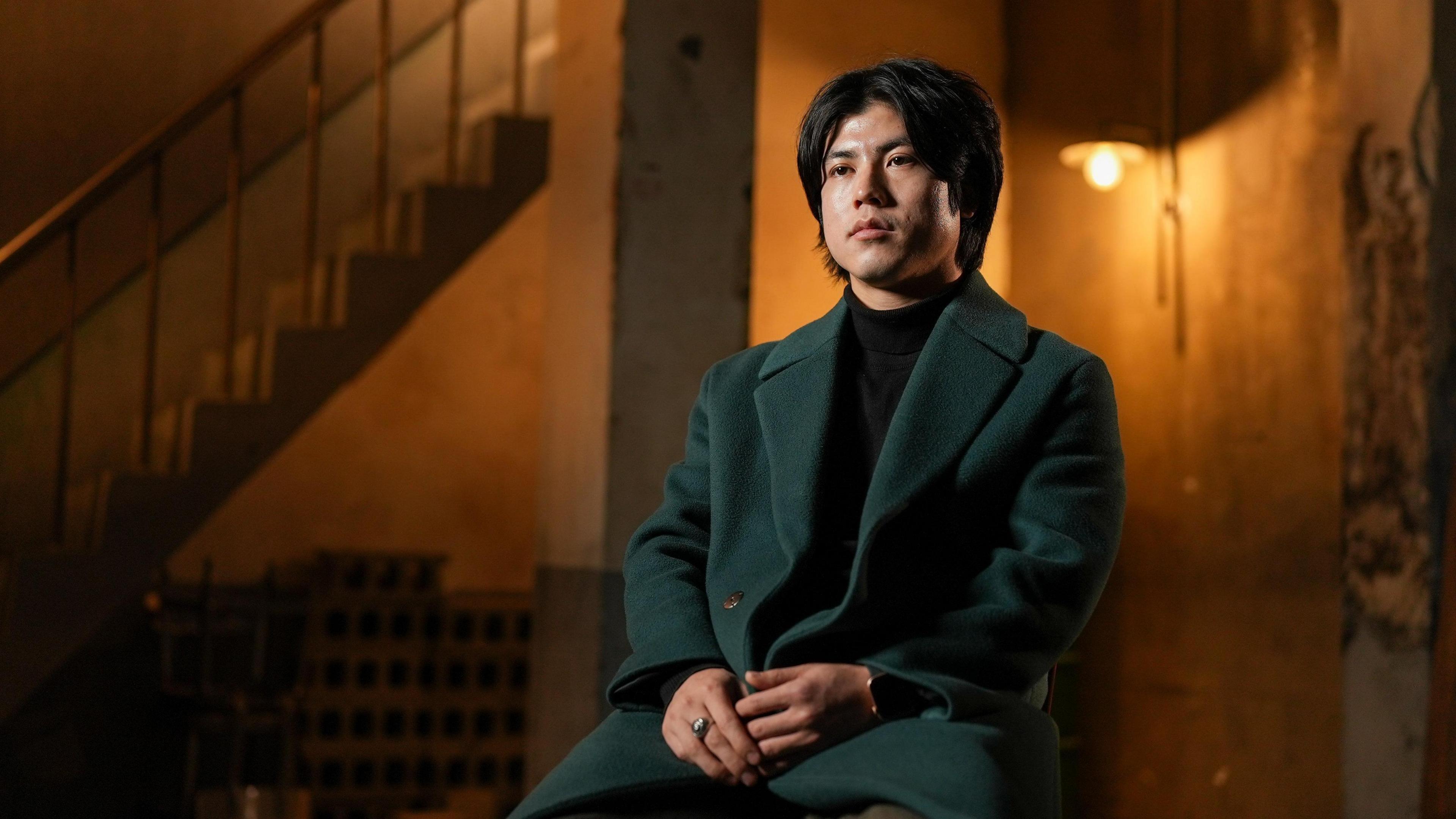 Haneul sitting on a chair in a dimly-lit room wearing a dark green coat while he speaks to the BBC.