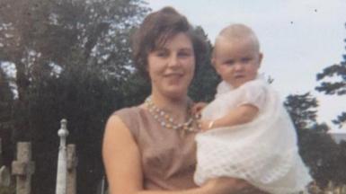 An old photo of Sandie Bowen as a young mum, holding her baby daughter Anita in her arms. Sandie is wearing a pink dress and colourful bead necklace and has short brown hair. Baby Anita is in a white Christening dress.