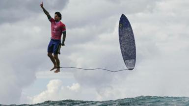 A surfer appears to be standing in the sky after being ejected from the sea. He has one finger raised and is pointing towards the sky. He wears a red T-shirt and blue shorts and his surfboard if pointing upwards too