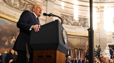 Donald Trump standing at the lectern speaking, with the paintings in the rotunda in the background