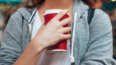 Woman wearing grey hoody holding a red disposable coffee cup close to her chest