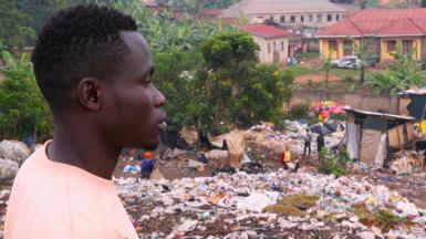 Okuku Prince in a pink T-shirt looking out over the Kiteezi rubbish dump in Kampala, Uganda, with houses seen in the background.