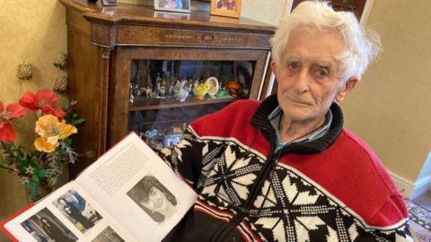 Dr Jim Swire sits at home holding a photo album which pictures his late daughter. He is wearing a red, white and blue jumper and has white hair. Behind him is a brown wooden unit with small figurines in, and there is a bouquet of artificial flowers with red and yellow petals to his left.