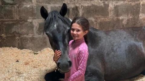 A girl of about 11 years is kneeling beside a black horse that is lying down. The horse's head is cradled in her hands and she is smiling. They are in a stable.