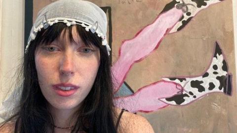 Poppy Baynham, a woman with long black hair and a lace napkin on her head standing in front of one of her paintings