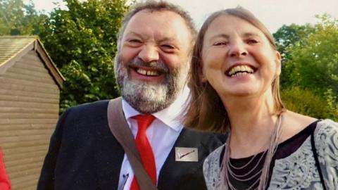 Richard Still and Lesley Clarke standing close together and smiling into the camera. Richard, who has short hair and a beard, is wearing a suit, white shirt and red tie. Lesley, who has shoulder length hair, is wearing a black top and a necklace.