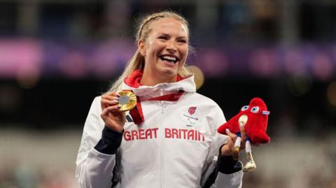 Wheelchair racer Samantha Kinghorn looks ecstatic as she holds up her newly-presented gold Olympic medal on the podium in Paris. She wears her Team GM tracksuit and beams at the appreciative crowd.