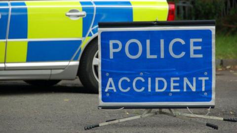 A blue sign says police accident on it. It is placed on a road with part of a police car showing in the background