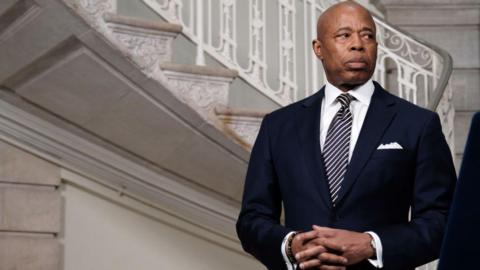 Eric Adams - a bald man - looks to the side, off camera. He is wearing a suit and there is a staircase behind him