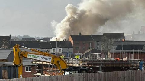 A large plume of smoke above a row of houses. There is a digger in the foreground and construction workers on a building site.