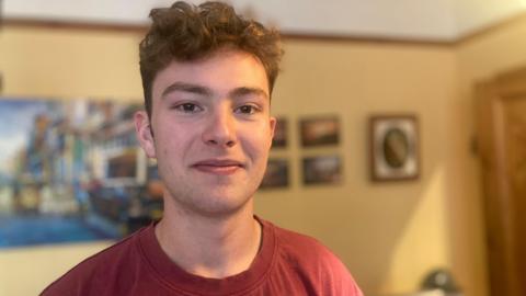 Manolo Niembro is smiling at the camera while wearing a red t-shirt. He has  brown eyes and light brown hair that is short on the sides and curly on top.  He is stood in a room with creamy yellow walls and framed pictures are behind him. 