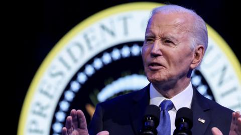 Joe Biden, dressed in a dark suit with a blue tie and white shirt, stands before a microphone, lifting his hands, in front of a seal that reads President of the United States.