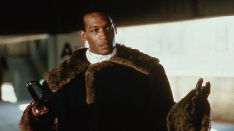 Screengrab of Tony Todd as his Candyman character in the 1992 film. He is wearing a fur coat and is stood with his arms semi-outstretched in front of him. He has a hook instead of a right hand, as per his character. His left hand has a glove on. He is clean shaven and has short black hair