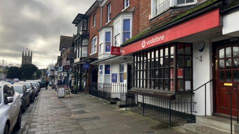 Red Vodafone banner above a shop doorway. The building has a large box window with a wooden frame and white plaster on the ground floor. The upper floor is brick and has a smaller white bay window.