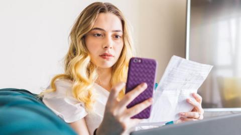 A young blonde woman with a look of concentration stares at her smartphone while holding a bill in her other hand