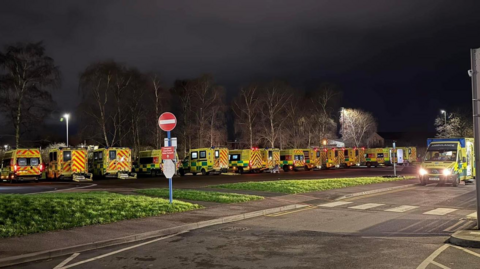 A row of ambulances lined up by a grass verge at night time. Another ambulance can be seen driving on a nearby road. 