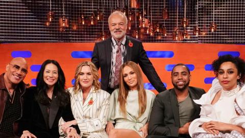 Graham Norton stands behind a sofa of guests.