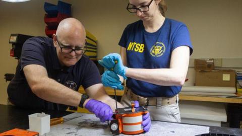 A man wearing purple latext gloves holds the orange casing of a black box, while an employee uses a tool to wedge open its casing.