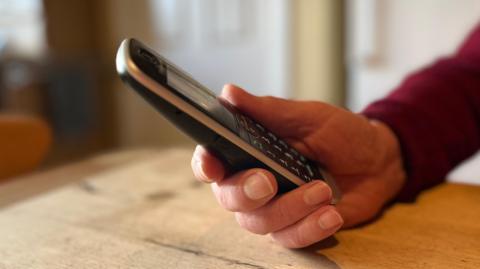 The man's hand holds his landline phone on a wooden table.