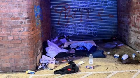 A sleeping bag and other items in a doorway with shutters covered in graffiti.