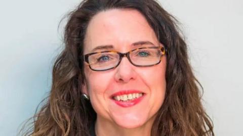 Close up headshot of Sharon Kemp with a white background. She is a white woman who appears to be in her late 40s or early 50s. She is smiling. She wears a little bit of makeup including red lipstick. She wears silver earrings. She has curly brown hair which is long enough to go out of shot.