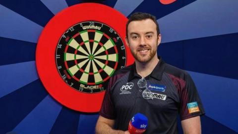 Huw Ware smiles looking at the camera. He is standing in front of a dart board and is holding a mic.