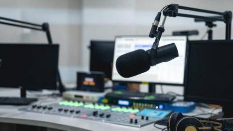 Image shows a radio desk including a microphone, sound keys and monitor. There are some headphones also sitting on the desk.