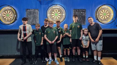 Members of Shooters Leominster Junior Darts Academy, wearing green club T-shifts, shorts and trainers, with darts boards behind them.