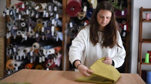 A woman wearing a white jumper working at a fabric shop in east London. She is folding some yellow material on a wooden counter, with rolls of material seen stacked behind her