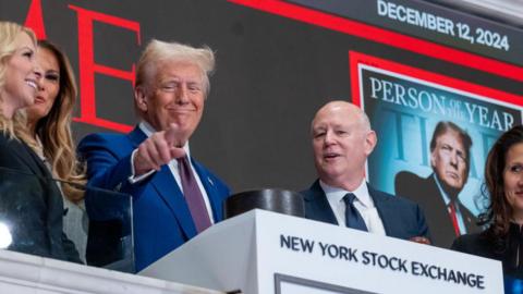 President-elect Donald Trump is seen standing above the New York Stock Exchange (NYSE) on December 12, 2024 in New York City with his wife, Melania, partially visible and a few others around him. He is pointing and a screen showing the Time magazine cover with him as Person of the Year is behind him. 