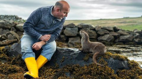 Billy Mail is sitting next to Molly the otter on rocks near the sea