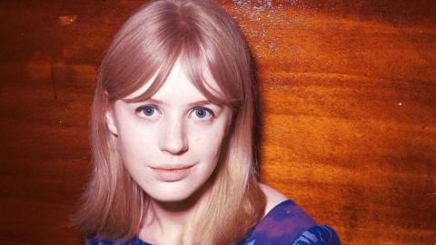 Marianne Faithfull in 1964. She is photographed above the shoulders against a polished wood background. She has light brown hair and a blue dress and is looking directly at the camera 