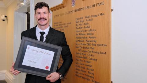 Boxer Nathan Heaney, holding a certificate and stood next to a wooden board listing Stoke-on-Trent's sporting stars.