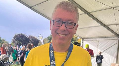 A head and shoulders picture of John Howard. He is wearing a yellow T-shirt with a medal around his neck, which says 'Great Eastern Run'. A crowd of people are behind him and a white tent is to the right. 