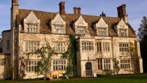 The outside of Anglesey Abbey. The sky is blue and the light is shining onto the building, casting it in a warm glow. The sand coloured stone is partly covered by ivy creeping up the walls of the Abbey. The is a wooden arched door in the centre of the building. 