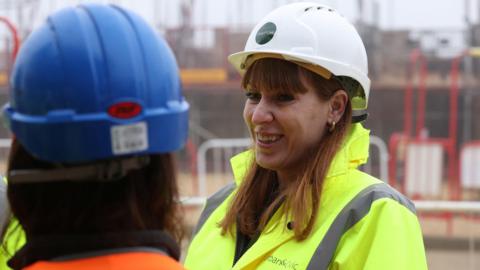 Angela Rayner wears a white hard hat and yellow hi-vis jacket. She is looking to the left of the camera and smiling at person in the foreground. 