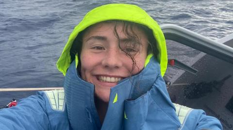 Zara Lachlan wearing a light blue coat with a neon yellow hood while on her boat in the middle of the ocean. She is smiling at the camera and a strand of her hair has come out from under her hood. It has fallen across her freckled face and is wet.  