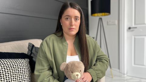 Victoria Buckley is wearing a white top and khaki green bomber jacket. She has long dark brown hair and is looking directly at the camera with a sad expression. She is holding a brown teddy bear that says "Ollie" on its jumper.