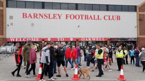 A crowd of people stand outside a building which has a sign which reads Barnsley Football Club on the side. 