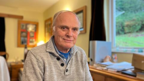 Ian Kell, an older man with grey hair and blue eyes, is looking at the camera. He is sitting at home, wearing a grey jumper with a blue shirt underneath