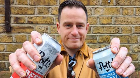 Ian Butt holding two cans of Potts' gravy. His arms are stretched out close to the camera. He is standing against a brick wall, looking directly at the camera and smiling. He has dark short hair and is wearing a mustard yellow jacket, with sunglasses hanging on his collar. 