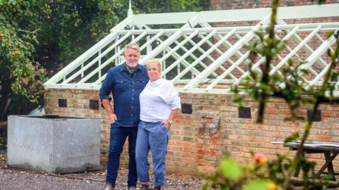 Jules and Mark standing in front of the Victorian greenhouse which they are restoring. Jules is wearing blue trousers and a white shirt with her hand in her pocket and her arm around Mark, who is standing to her left wearing blue jeans and a denim shirt. Behind them is a low brick greenhouse with new white roof rafters, but no glass panels fitted yet. The couple are looking at the camera and smiling.