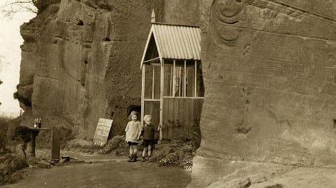 Two children by a wooden porch with a corrugated iron roof, next to a rock face. They have shorts on and are seen in sepia print. Behind them is a sign with a chalk message on it and nearby a wooden rough table with bottles on.