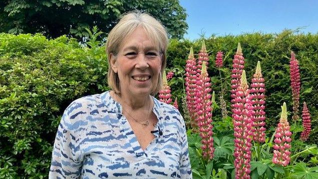 Lynda Sawyer smiling into the camera as she stands in front of a high green hedge and tall pink flowers. She has blonde hair cut into a bob and is wearing a blue and white patterned blouse.