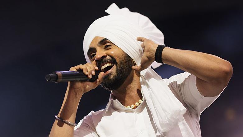 Singer Diljit Dosanjh wearing all white traditional Punjabi attire including a white Turban (religious headwear), while singing into a black microphone, with his left hand touching his head. The background is dark with a spotlight.