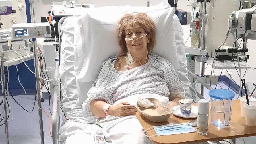 Lynda Sawyer wearing a hospital gown and propped up by pillows in a hospital bed surrounded by medical equipment. She is wired up to monitors and has a nasal cannula for oxygen. There are various items on the wooden tray positioned over one side of the bed, including a water jug and cups.