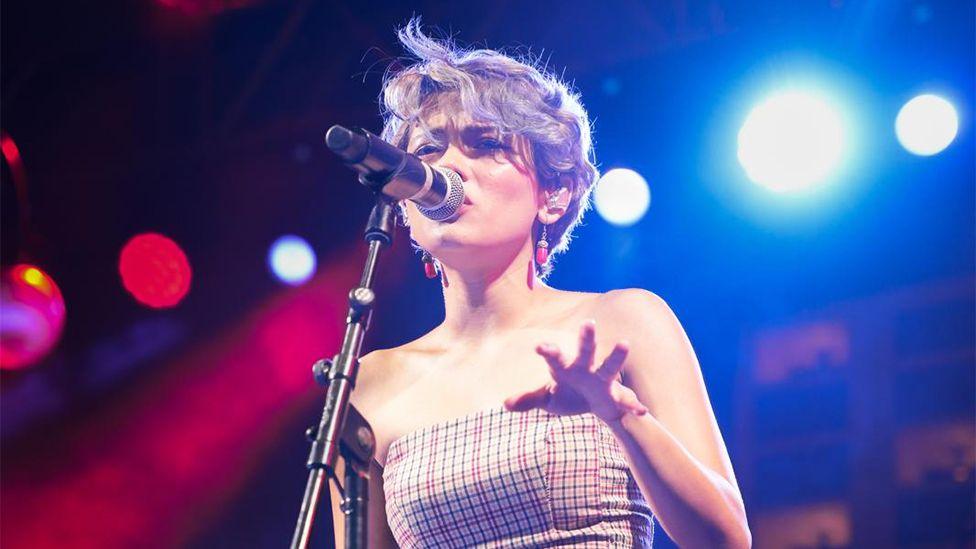 Anoushka singing into a microphone, with blue, white and red spotlights behind her. Her left hand is pointing out towards the crowd.