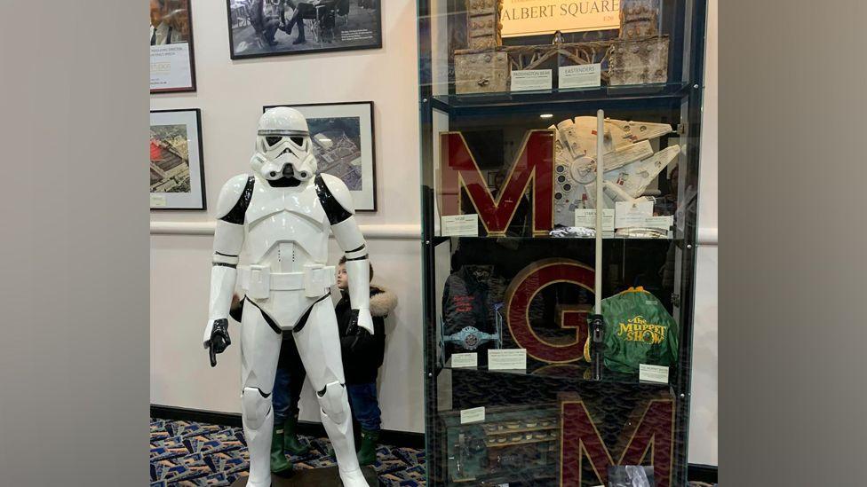 The exhibition at the cinema. Framed photos on the wall sit next to a glass cabinet holding various items, including an Albert Square Sign, the MGM studio tower letters, a Muppet Show bag, and a Star Wars Millenium Falcon. A full sized Storm Tropper model stands next to the cabinet and there is a child behind it.